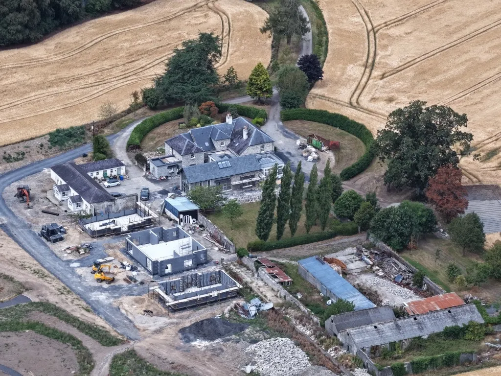 Aerial view of a stone house under redevelopment next to a new-build structure under construction, surrounded by farmland